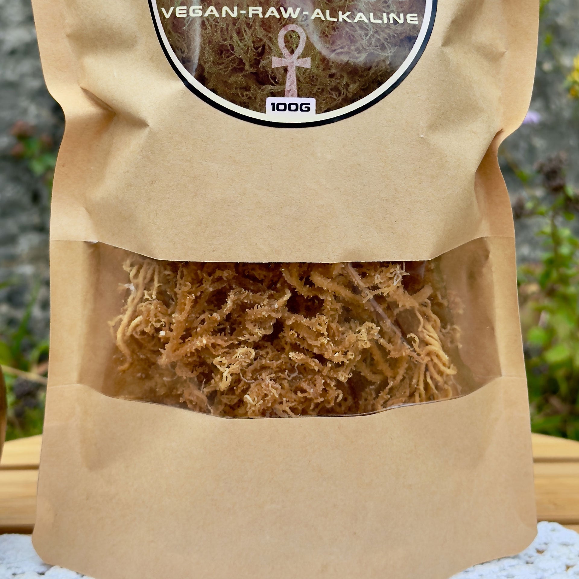 Nahaufnahme der ZeaMoss Raw 100g Packung mit Goldenem Sea Moss, auf einem Tisch in natürlicher Umgebung platziert. Im Hintergrund grüne Pflanzen und eine Steinwand
