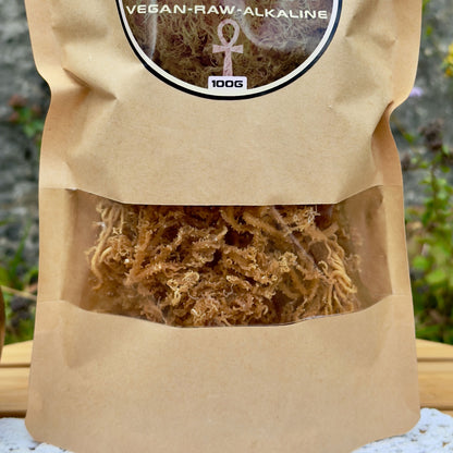 Nahaufnahme der ZeaMoss Raw 100g Packung mit Goldenem Sea Moss, auf einem Tisch in natürlicher Umgebung platziert. Im Hintergrund grüne Pflanzen und eine Steinwand