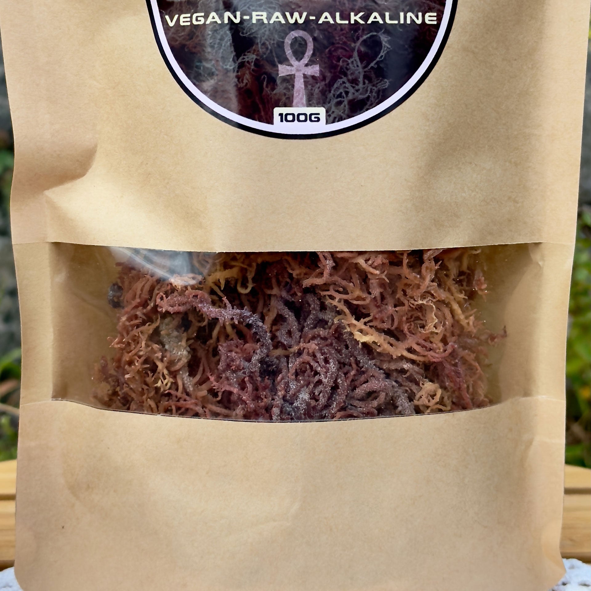 Nahaufnahme der ZeaMoss Raw 100g Packung mit Rainbow Sea Moss, auf einem Tisch in natürlicher Umgebung platziert. Im Hintergrund grüne Pflanzen und eine Steinwand