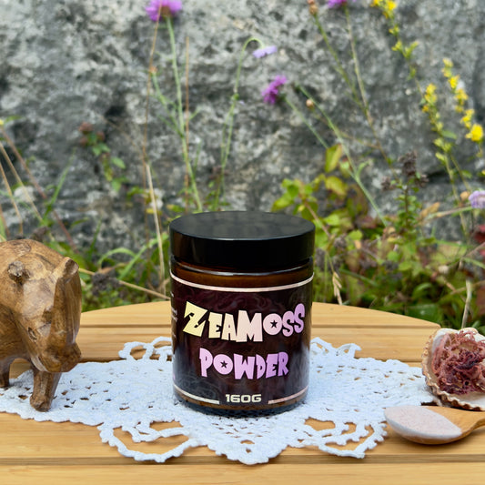 ZeaMoss Powder 160g Wildcrafted Rainbow Sea Moss (Euchema Denticulatum) aus East Africa, Tansania, präsentiert draußen vor einer Naturkulisse mit Blumen, Dekoration mit Holznashorn, Muschel mit Rohalge und Holzlöffel mit Pulver
